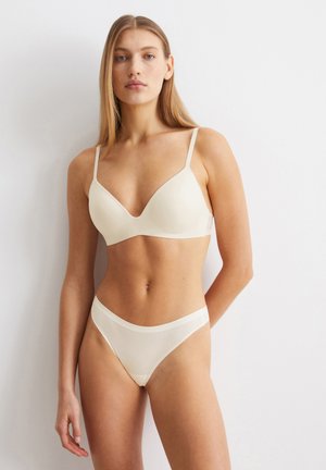 Reggiseno beige in morbido tessuto liscio, con scollatura a V profonda e spalline regolabili; bikini coordinato in rete trasparente con vita elastica e copertura minimale.