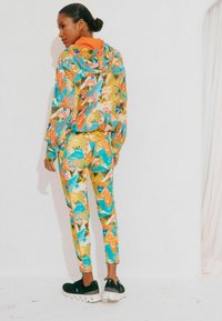 Tuta colorata con un mix di motivi arancioni, blu verde e gialli. La giacca con cappuccio ha maniche lunghe; i pantaloni sono aderenti. Sneakers nere.