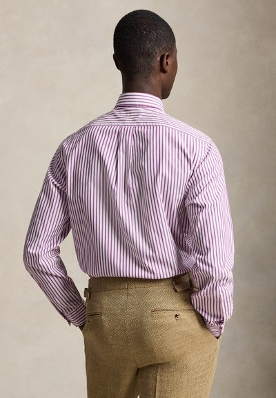 Homme de dos portant une chemise à manches longues rayée rose et blanche rentrée dans un pantalon marron texturé, les mains dans les poches.