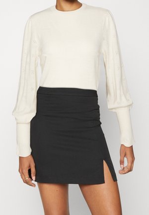 Kvinde iført en hvid langærmet strikket sweater med pufærmer og en sort mininederdel med slids foran.