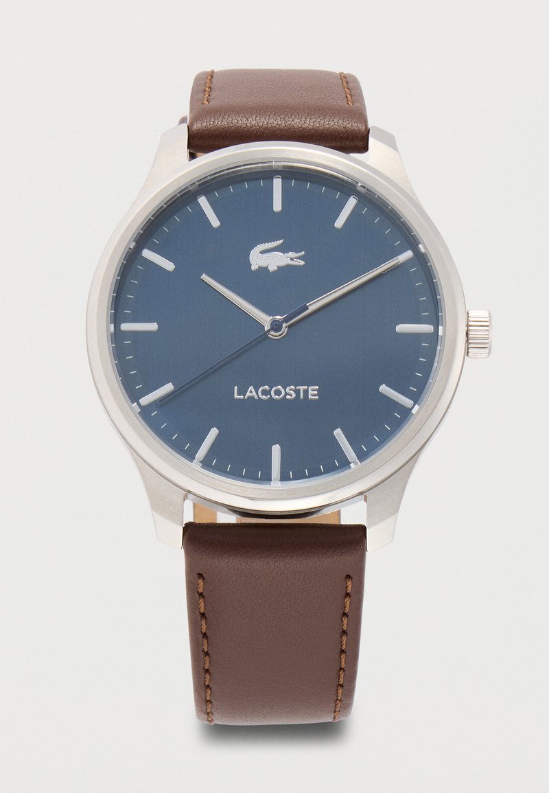 Στρογγυλό ασημί ρολόι χειρός με μπλε καντράν, ασημένιους δείκτες ώρας και δείκτες, καφέ δερμάτινο λουράκι, λογότυπο και όνομα της μάρκας Lacoste στο πρόσωπο.