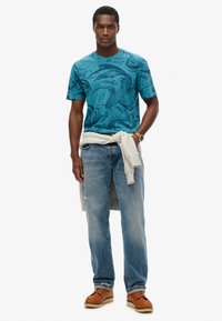 Camiseta gráfica azul con patrones de olas y peces, combinada con jeans azul desgastados y un suéter gris claro atado a la cintura. Zapatos marrones.