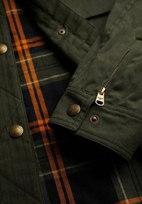 Chaqueta acolchada verde oliva con botones y cremallera de latón; tela a cuadros con rayas en azul marino, naranja y blanco visibles por debajo.