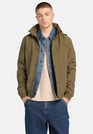 Junger Mann mit kurzen Haaren, der eine khakifarbene Jacke übereinander, eine blaue Jeansjacke, ein beiges Hemd und blaue Jeans trägt, mit den Händen in den Taschen, vor weißem Hintergrund.