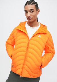 Padded oransje jakke med horisontal quilting, tettsittende hette og glidelås. Har logo på venstre bryst og glidelåser på lommene.