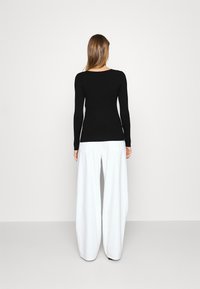 Femme aux longs cheveux se tient de dos, portant un haut noir ajusté à manches longues et un pantalon blanc large et décontracté devant un fond uni.