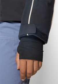 Manche d'une veste noire avec une bordure blanche et une fermeture Velcro, superposée à une manche noire ajustée. Main visible portant une chemise bleue.
