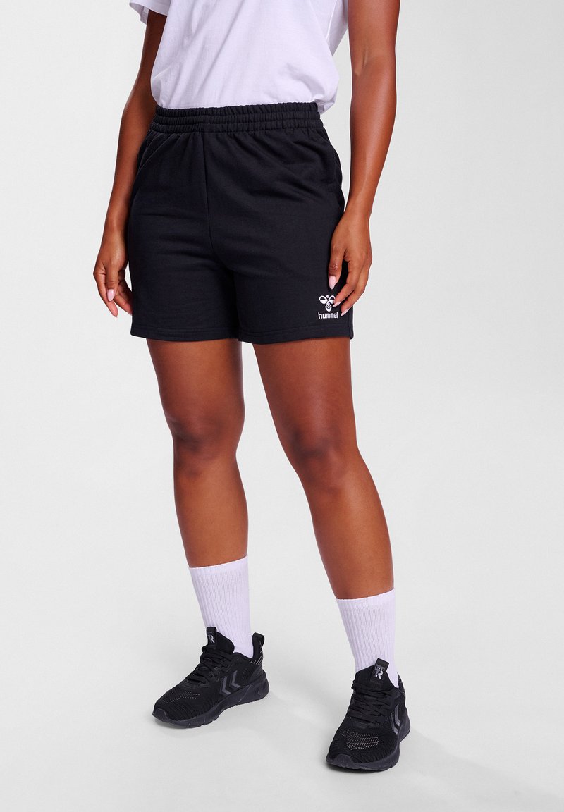 Svart bomullsshorts med elastisk midje og sidelommer, som har en hvit logo på venstre ben. Brukt med svarte sneakers og hvite sokker.