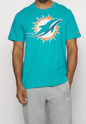 Mann trägt ein türkisfarbenes Miami Dolphins T-Shirt mit Delfin-Sonnen-Logo, kombiniert mit hellgrauen Nike-Sweatpants, steht mit einer Hand in der Tasche.
