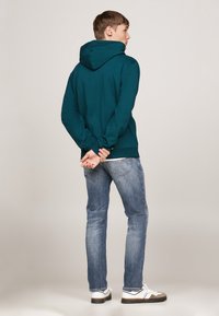 Sudadera en color verde azulado con una gran capucha, mangas largas y dobladillo acanalado, combinada con jeans azul claro y zapatillas blancas con acentos oscuros.