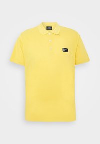 Polo shirt jaune à manches courtes avec trois boutons blancs et un petit patch logo rectangulaire noir sur la poitrine gauche, sur fond blanc.