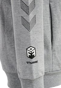 Felpa grigia con grafiche a chevron nere sulle maniche, un badge con logo sul polsino e un tessuto morbido e strutturato.