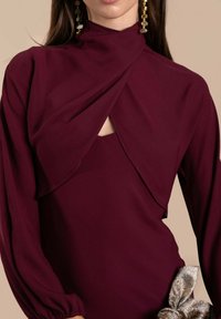 Bordeauxfarbenes Kleid mit drapiertem Oberteil, hohem Kragen und langen Puffärmeln. Mit einem Ausschnitt am Hals und einem metallischen Schleifenelement in der Taille.