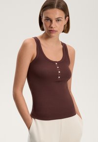 Rjava rebrasta majica brez rokavov s kroženim izrezom, ki ima gumbasto zapenjanje spredaj in gladko teksturo. V kombinaciji s svetlimi hlačami.