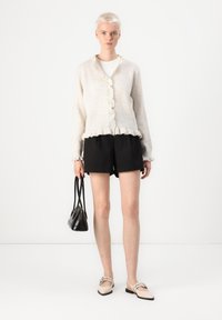 Cardigan in lana beige con orlo a coste e bordo arricciato, con bottoni sulla parte anteriore. Abbinato a pantaloni corti neri e una piccola borsa nera.