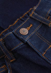 Gros plan sur un tissu denim bleu foncé avec des surpiqûres orange, un bouton en métal orné d'une étoile, et des détails de passants de ceinture.