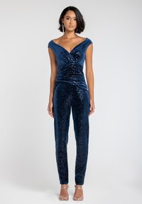 Jumpsuit blu navy con paillettes, design off-the-shoulder, fronte plissettato e silhouette aderente. Texture liscia e dettagli lucidi. Sfondo chiaro.