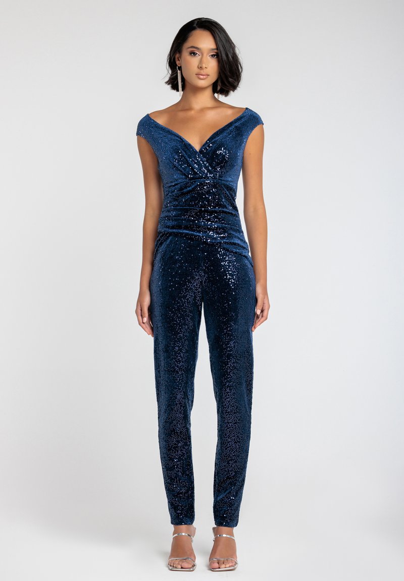 Jumpsuit blu navy con paillettes, design off-the-shoulder, fronte plissettato e silhouette aderente. Texture liscia e dettagli lucidi. Sfondo chiaro.