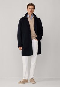 Giovane uomo vestito con un soprabito nero, maglione beige sopra una camicia a righe blu, pantaloni bianchi e scarpe beige con lacci, in piedi contro uno sfondo semplice.