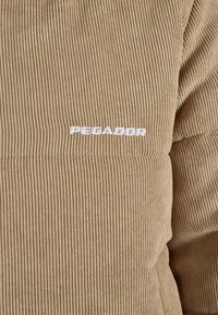 Kremas spalvos šonkaulinė medžiaga su minkšta tekstūra, su balta "PEGADOR" užrašu. Medžiaga atrodo šilta ir neformali.