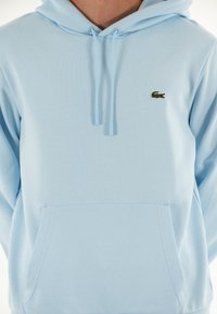 Sudadera azul clara hecha de algodón suave, con un bolsillo tipo canguro y cordones ajustables. Incluye un pequeño logotipo bordado en el pecho.