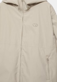 Bézs kapucnis dzseki elülső cipzárral, kis hímzett logóval a mellrészen és cipzáras zsebbel az alsó oldalon.
