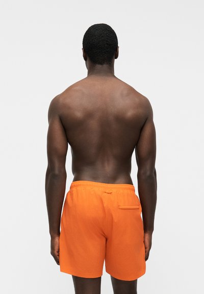 Homme à la peau foncée et aux cheveux courts et bouclés, debout de dos, portant un short de bain orange vif sur fond blanc uni.
