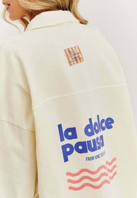 Krémová oversize bunda s modrým grafickým textom "la dolce pausa" a červeným vlnovým dizajnom, pričom na zadnej strane sa nachádza etiketa so prúžkovaným vzorom.