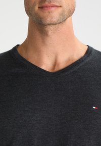 Pull à col en V gris foncé en tissu tricoté, avec un petit logo rouge, blanc et bleu sur le côté gauche de la poitrine.