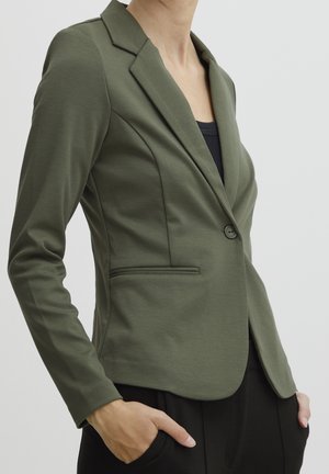 Kobieta w dopasowanej oliwkowo-zielonej marynarce na jeden guzik i czarnych spodniach, z rękami w kieszeniach, na jednolitym tle.