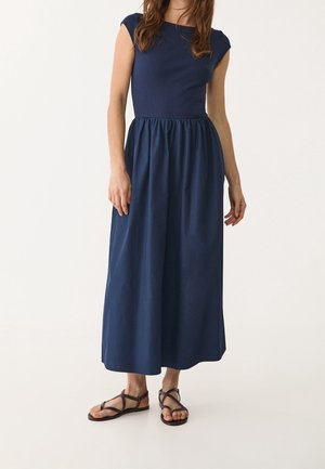 Vrouw draagt een navyblauwe maxi-jurk zonder mouwen met een geplooide taille en zwarte sandalen met bandjes en platte zolen, staand tegen een effen achtergrond.