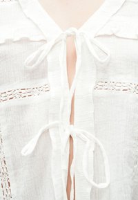 Λευκή μπλούζα με στρογγυλή λαιμόκοψη με βολάν και κλείσιμο με κορδόνι. Διαθέτει υφή υφάσματος και περίπλοκες δαντελένιες ενθέσεις σε κάθε πλευρά του ανοίγματος.