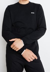 Pull noir avec un col rond, des épaules côtelées et des poignets blancs contrastants. Présente un petit logo "BOSS" sur la poitrine.