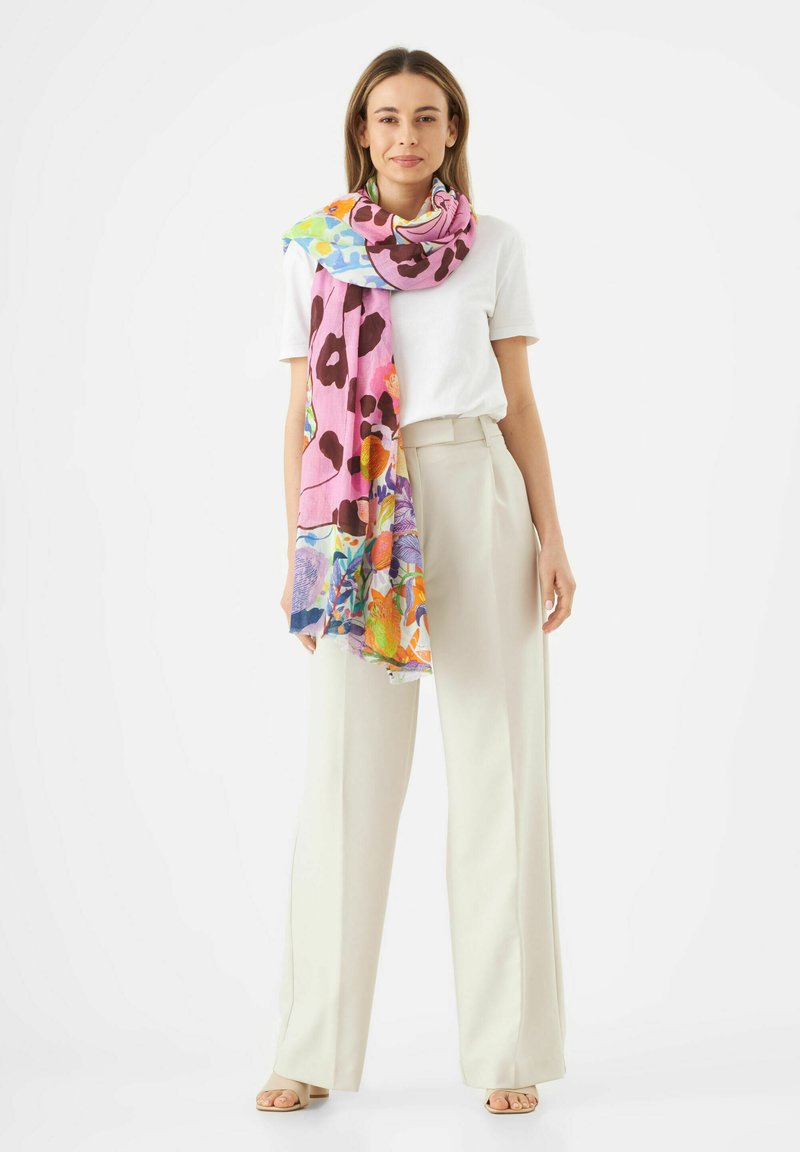 Écharpe légère à motif fleuri en rose, avec des accents orange, vert et bleu, drapée sur un haut blanc, portée avec un pantalon beige.
