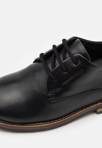 Chaussure en cuir noire à bout arrondi, avec quatre œillets pour les lacets, des détails de couture visibles et une semelle de couleur claire. Texture lisse avec des accents minimes.
