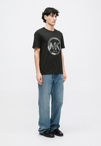 T-shirt preta de algodão com um logótipo cinza circular com o texto "MK" e "MICHAEL KORS". Modelo veste jeans azuis e sapatos pretos.