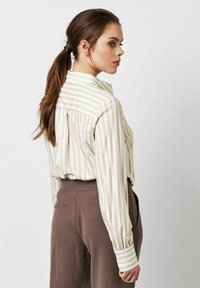 Striped long-sleeve blouse in cream and light brown, featuring a button collar and pleated back, paired with brown high-waisted trousers.