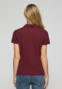 Burgundy pikétröja med krage, korta ärmar och en mjuk textur; kombinerad med ljusblå jeans med synliga fickor bak.