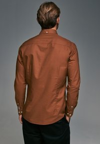 Camisa de manga larga marrón con costura en el yugo trasero, puños abotonados y dobladillo redondeado. Textura suave con un brillo sutil.