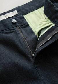 Dark denim pants with a green inner lining, zipper fly, and button closure. Features a smooth texture and tailored design for regular fit.