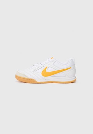 Chaussure de sport blanche avec un swoosh Nike jaune et une semelle couleur gomme, vue de côté sur un fond gris uni.