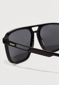 Μαύρα γυαλιά ηλίου με σκούρους φακούς, με γεωμετρικό σχέδιο πλαισίου και εγγεγραμμένο λογότυπο "HUGO" στο βραχίονα. Κόκκινες λεπτομέρειες στους βραχίονες.