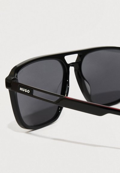 Lunettes de soleil noires avec des verres foncés, présentant un design de monture géométrique et un logo "HUGO" gravé sur la branche. Détails rouges sur les branches.