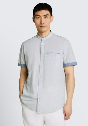 Man draagt een lichtblauw, kort mouw, overhemd met mandarijnenkraag en blauwe bies, gecombineerd met een witte broek, staand tegen een effen achtergrond.