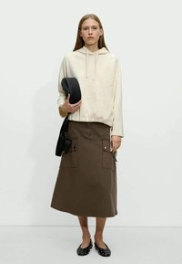 Giovane donna in felpa color crema, gonna midi marrone con tasche frontali, ballerine nere con borchie, che tiene una borsa rotonda nera e indossa una borsa a tracolla.