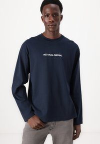 Camiseta de manga larga color marino hecha de algodón, con un logo de texto blanco "RED BULL RACING" centrado en el pecho. Modelo vistiendo pantalones grises.