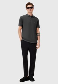 Dunkelgraues Poloshirt mit Kragen, zwei Knöpfen und kleinem Logo; kombiniert mit schwarzen Hosen und schwarzen Schuhen. Zu den Accessoires gehören Sonnenbrillen.