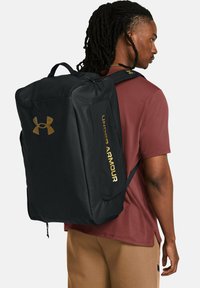 Zaino nero con logo dorato "Under Armour", design rettangolare, texture liscia, spallacci regolabili e una tasca frontale con zip.