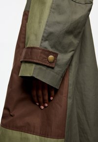 Chaqueta con bloques de color que presenta paneles en verde, marrón y oliva. El puño incluye un botón dorado y costuras detalladas. Tejido suave y texturizado.