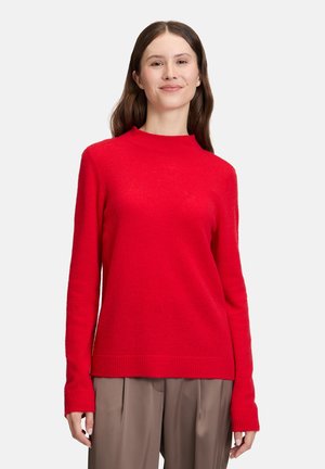 Jonge vrouw met lang bruin haar, gekleed in een rode trui met lange mouwen en taupe wijde broek, staand tegen een effen achtergrond.